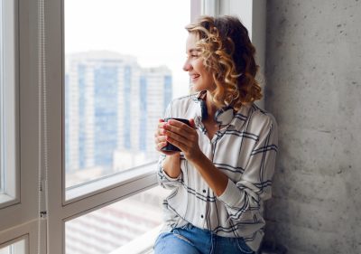 Dreamy wonder  woman looking into the window  , holding cup of hot  tee.  Grey urban wall  background.