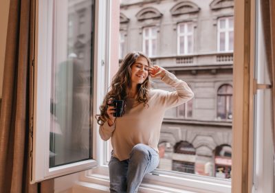 Refined caucasian girl in trendy shirt enjoying city views from window. Attractive curly woman chilling at home, sitting on sill with smile.